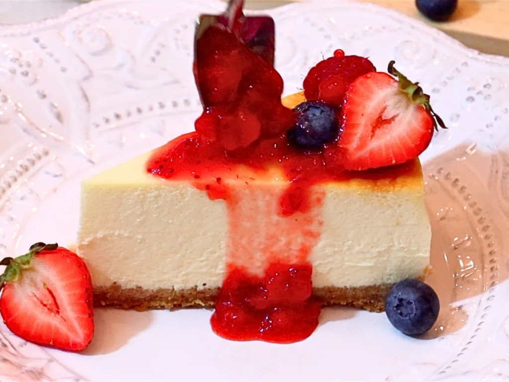 a slice of baked cheesecake with homemade strawberry sauce and fresh fruits on top